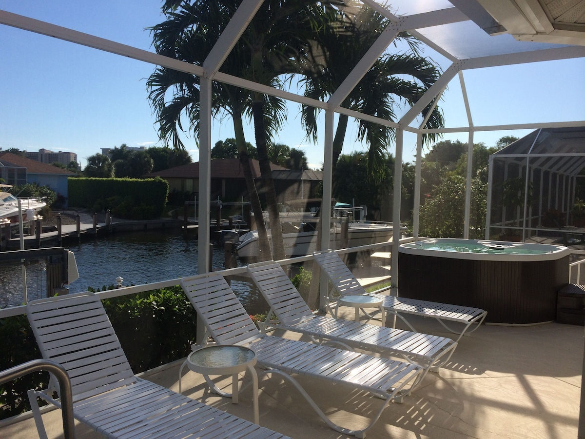 A covered outdoor area with lounge chairs and a hot tub, overlooking a body of water and palm trees.