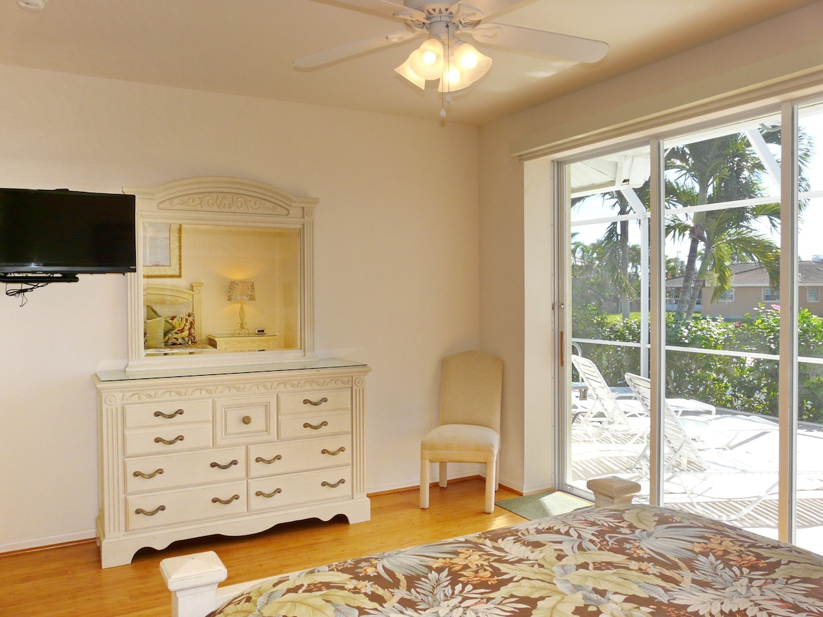 A bedroom with a bed, a dresser, a chair, a ceiling fan, and a TV mounted on the wall.