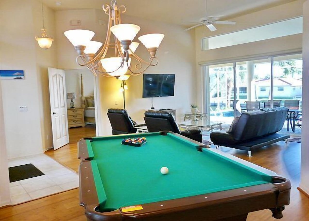 A pool table with a green felt surface, a white cue ball, and a set of pool cues. There is a chandelier with multiple light fixtures above the table.