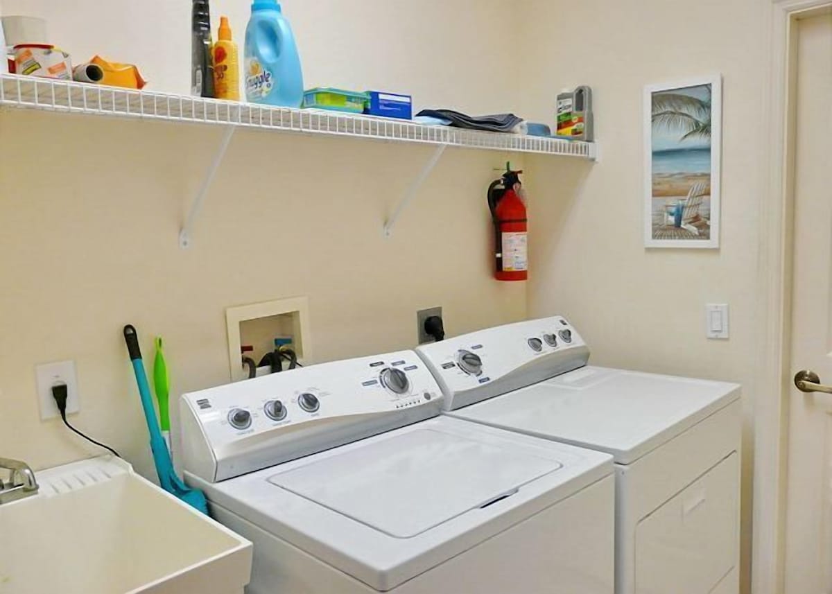 A laundry room with two front-loading washers and dryers, a shelf with cleaning supplies, a fire extinguisher, and a framed picture on the wall.