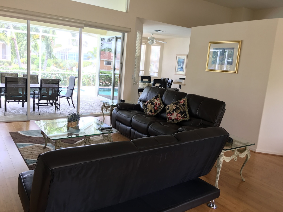 A living room with a black leather sofa, glass coffee table, and a view of a patio with outdoor furniture and a pool.