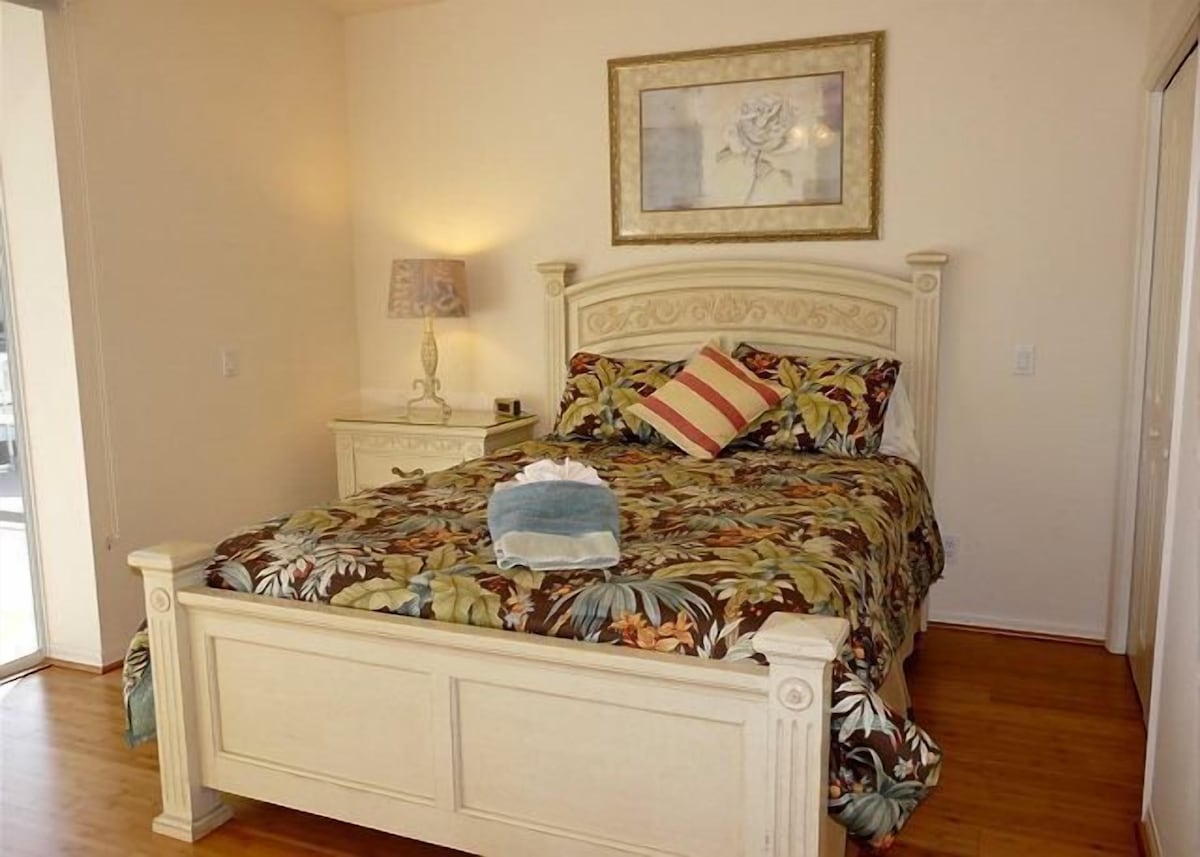 A bedroom with a floral-patterned bedspread, a single pillow, a bedside table with a lamp, and a framed picture on the wall.