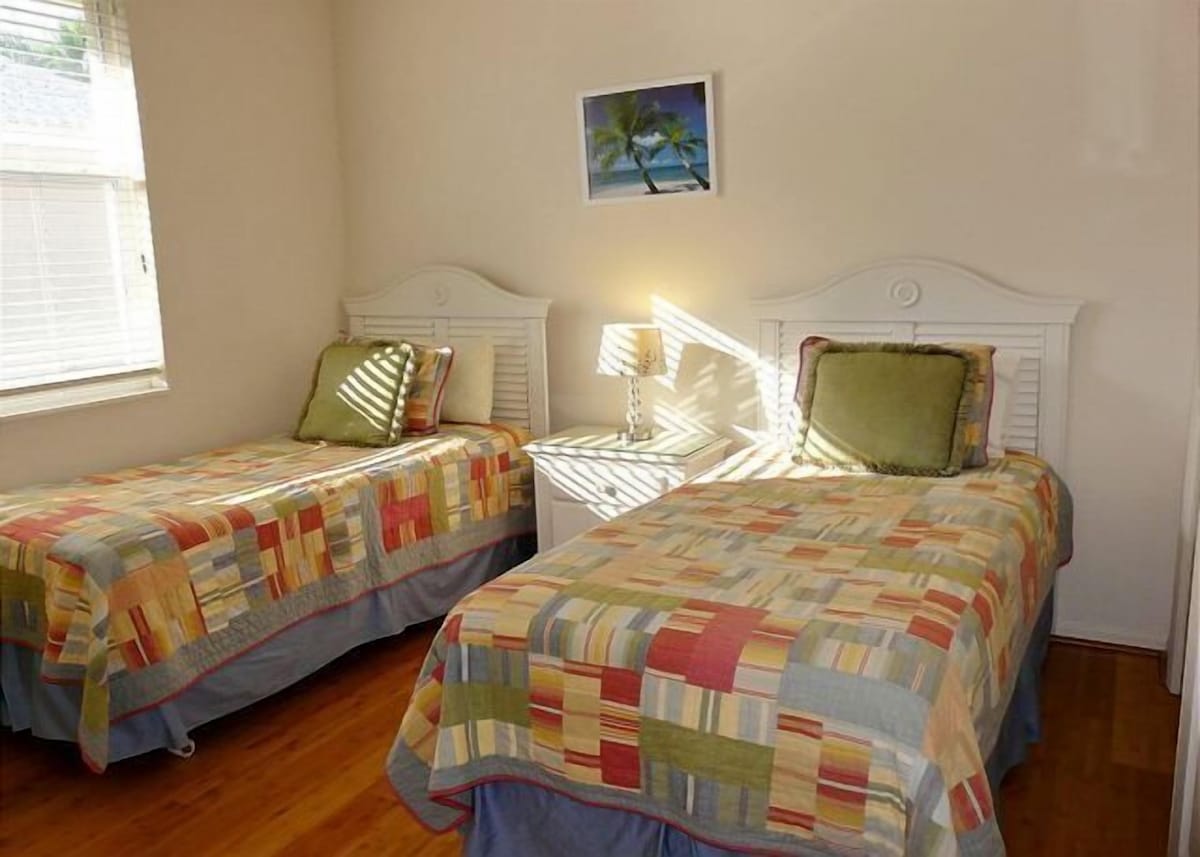 Two single beds with colorful quilts, a nightstand with a lamp, a small painting on the wall, and a window with blinds.