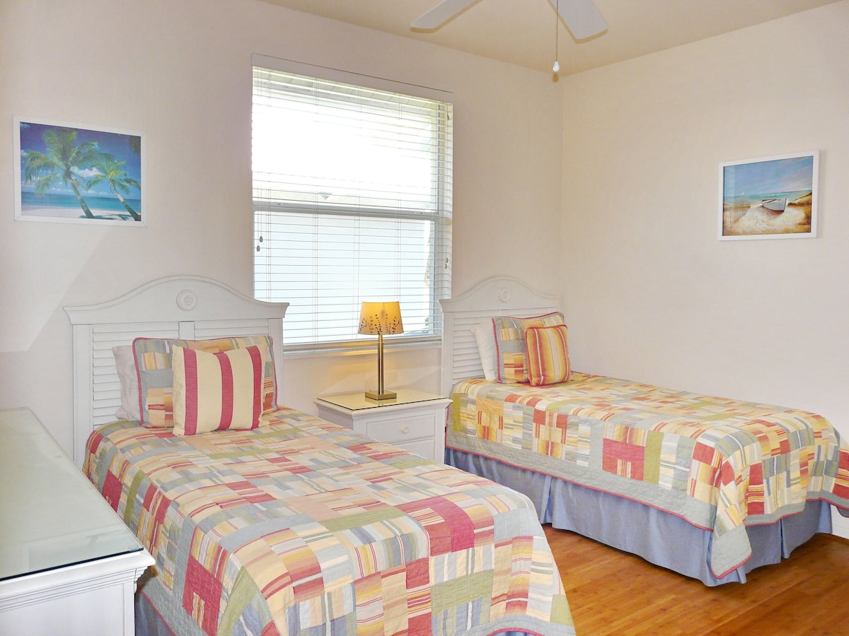 A bedroom with two single beds, a nightstand with a lamp, a dresser, and a window with blinds.