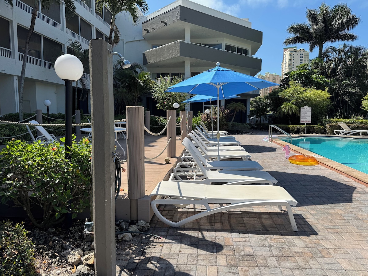 A poolside area with lounge chairs, a blue umbrella, and a sign indicating pool rules.