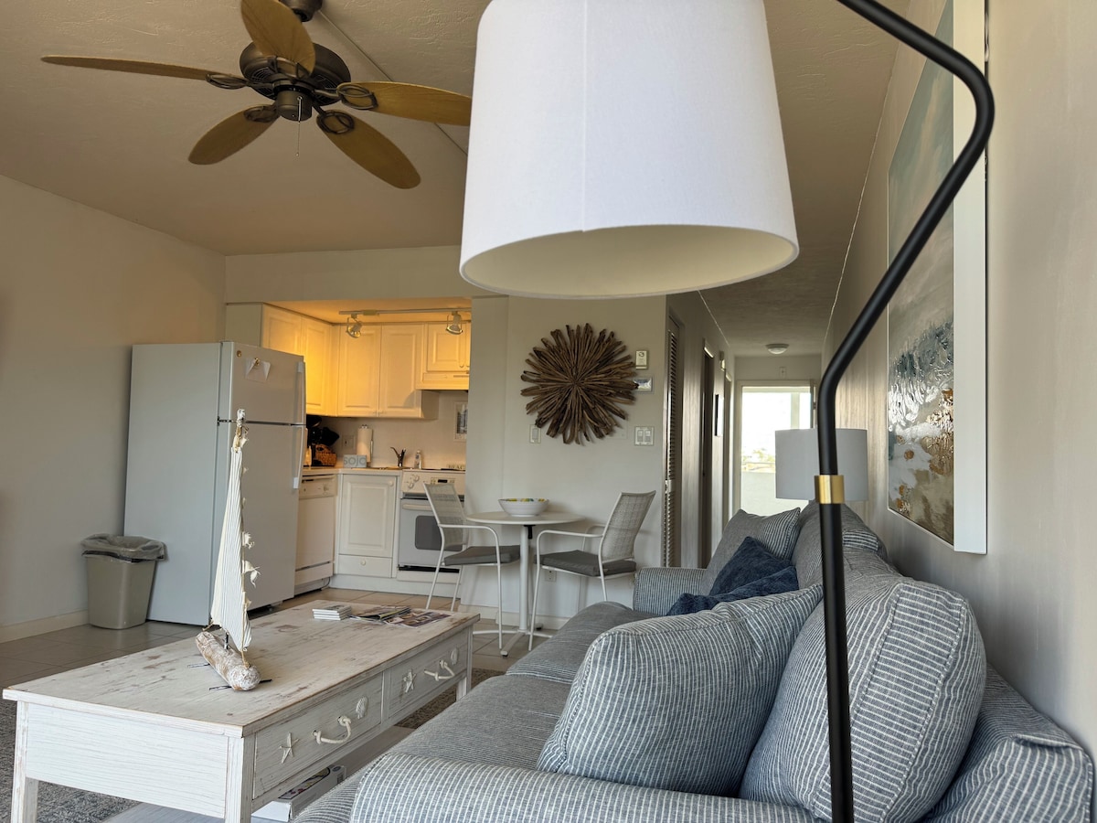 A modern living room with a sofa, a coffee table, and a ceiling fan. There is a kitchen visible in the background with white cabinets and a dining area with a table and chairs.