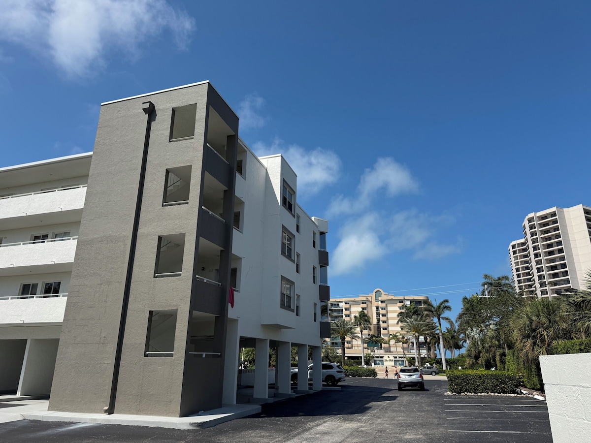 A modern multi-story building with balconies, a parking area, and palm trees in the background.