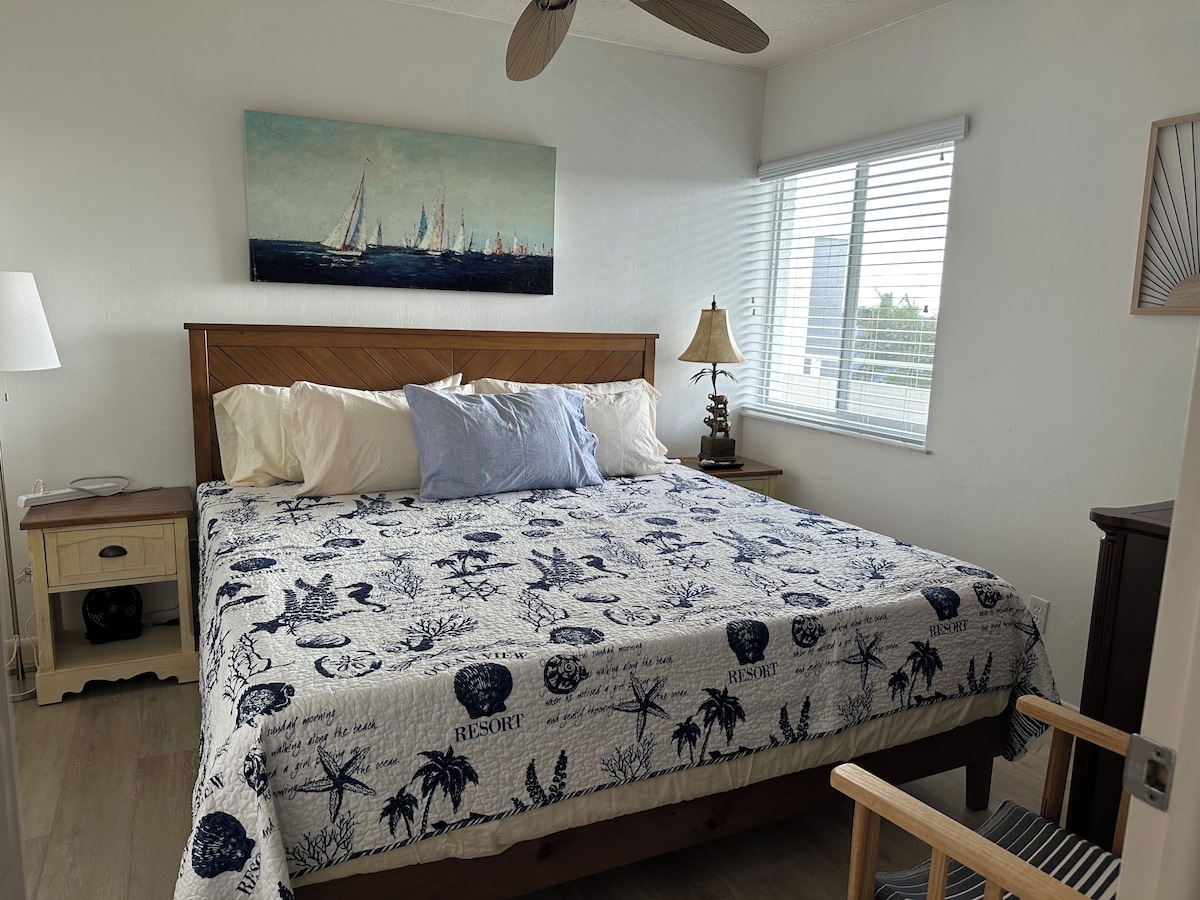 A bedroom with a bed, nightstand, lamp, and a painting of sailboats.