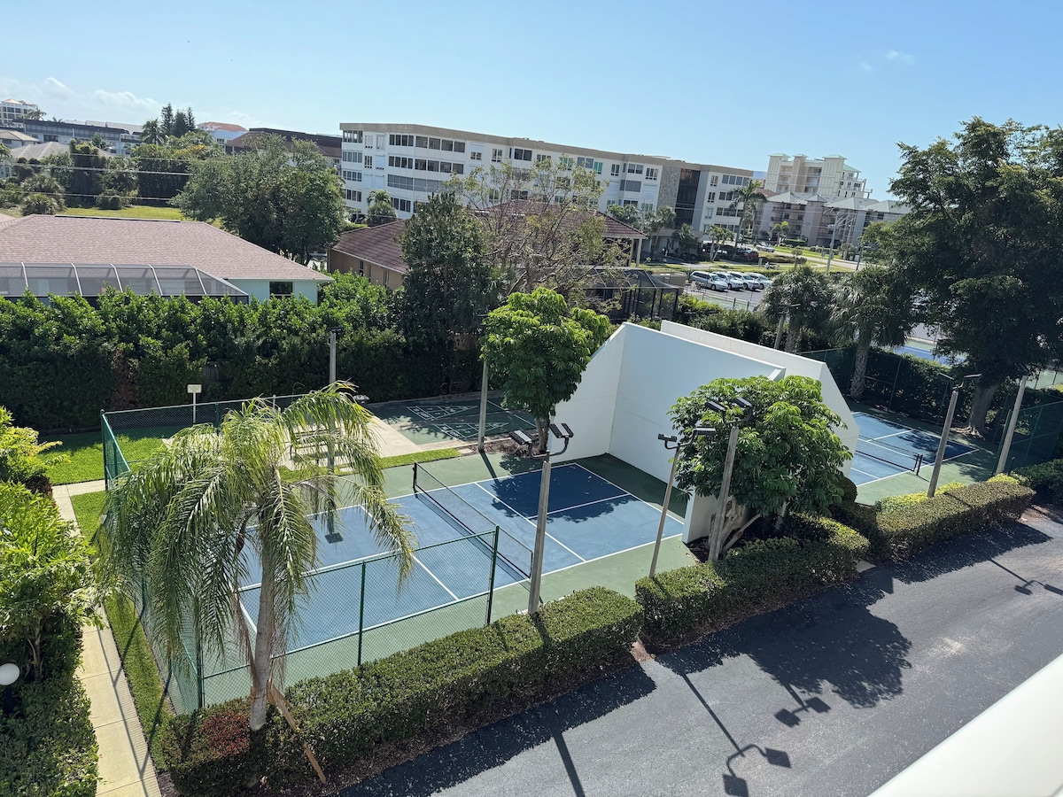 A tennis court surrounded by greenery and buildings.