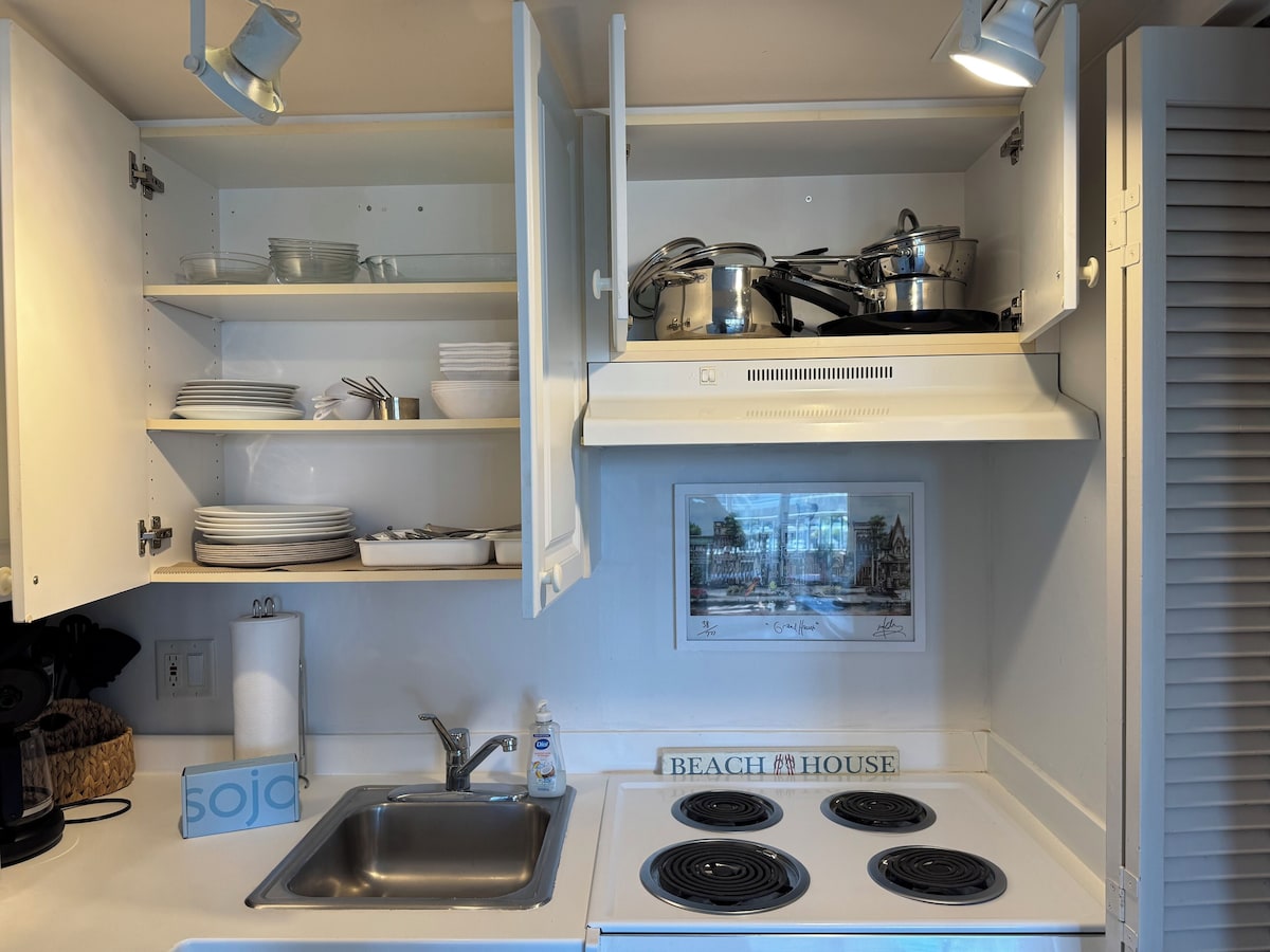 A compact kitchen with a sink, stove, and shelves containing dishes and cookware.