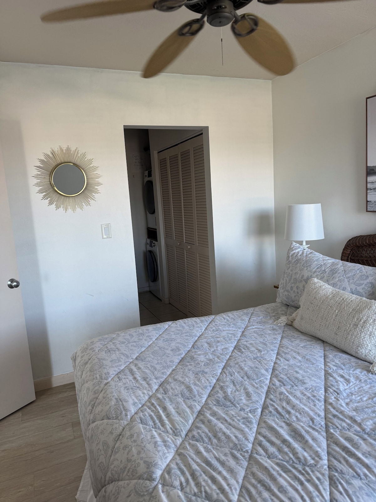 A bedroom with a bed, a ceiling fan, a wall-mounted mirror, a door, and a bedside lamp.