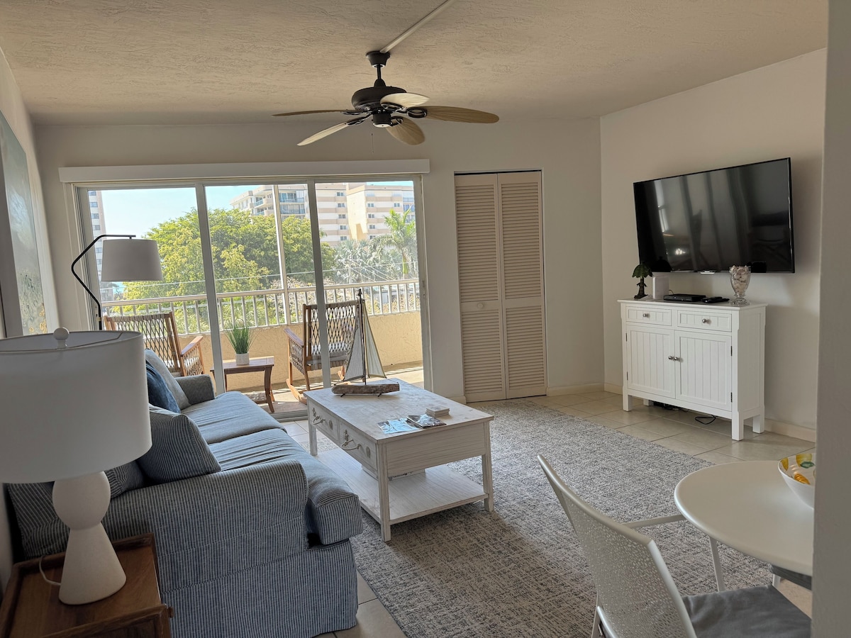 A living room with a sofa, coffee table, TV, and a balcony with a view of buildings and trees.