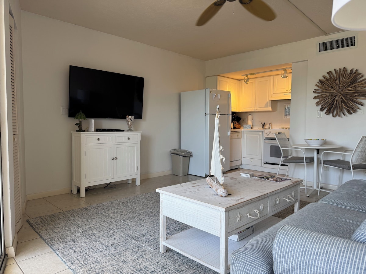 A compact living space with a flat-screen TV, a white cabinet, a dining table with chairs, and a kitchenette in the background.