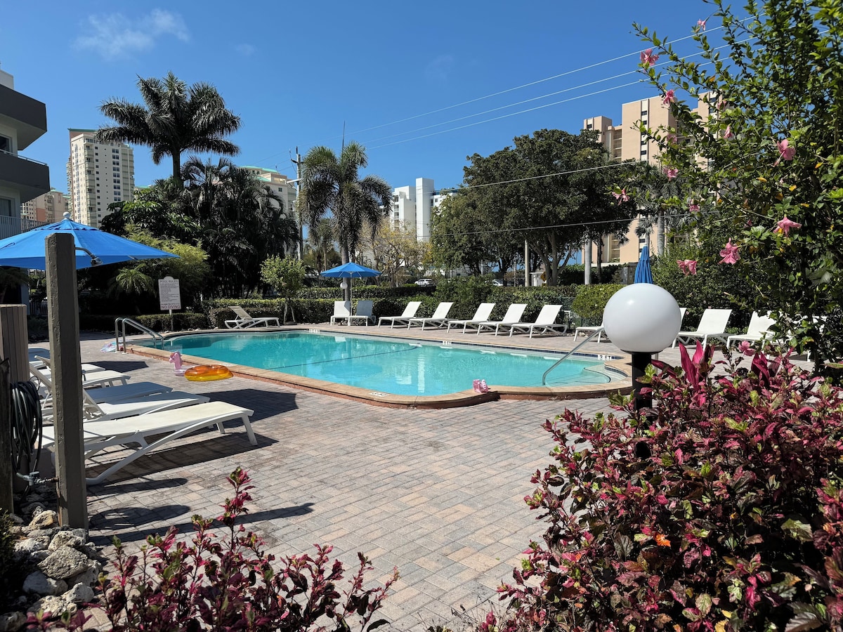 A swimming pool with lounge chairs and umbrellas, surrounded by palm trees and buildings.