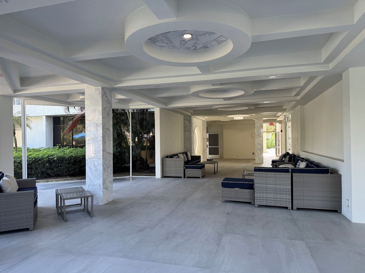 A spacious indoor area with a modern ceiling design, a seating area with sofas and chairs, and a view of greenery outside.