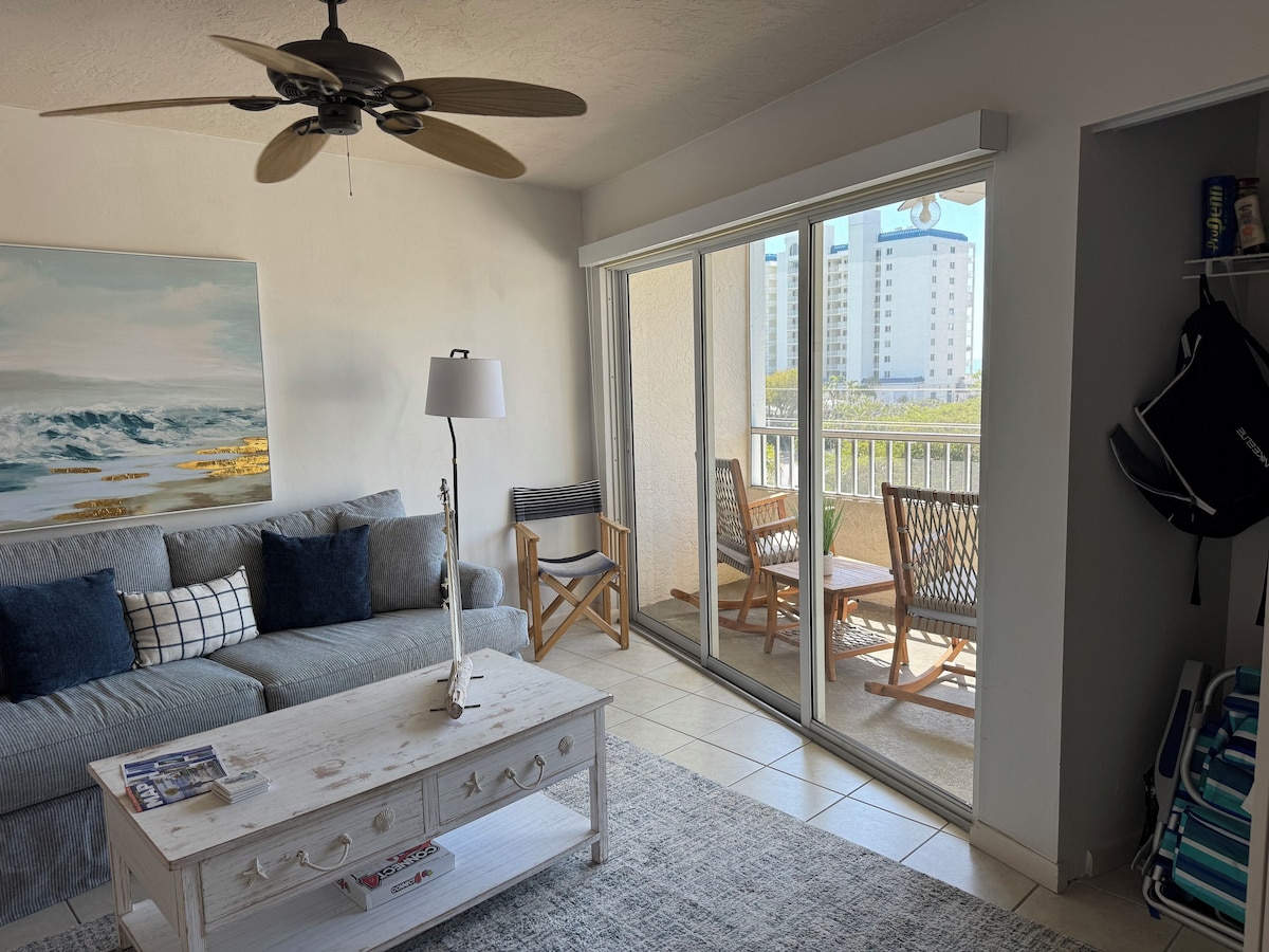 A living room with a sofa, a coffee table, and a floor lamp. There is a balcony with a view of buildings.