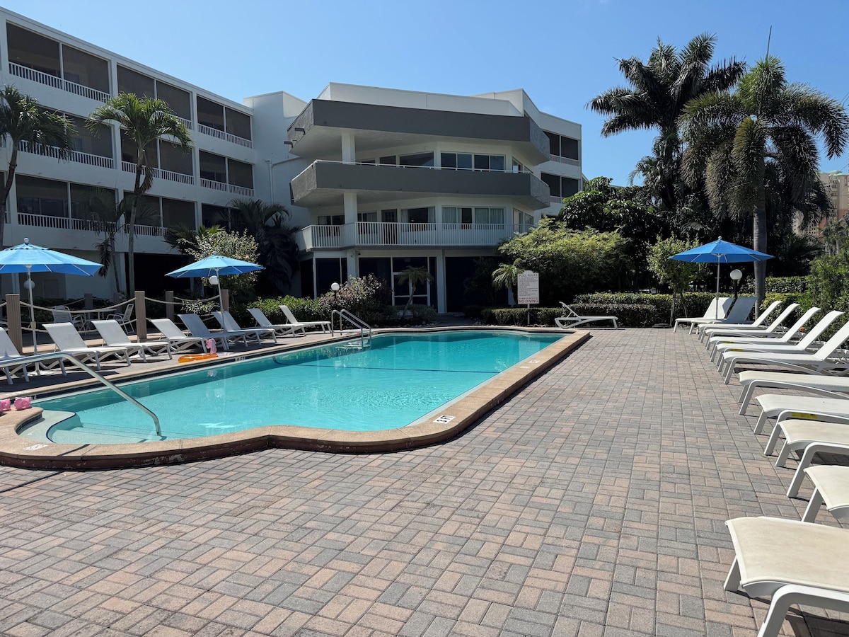 A swimming pool with lounge chairs and umbrellas, surrounded by a brick walkway and palm trees.