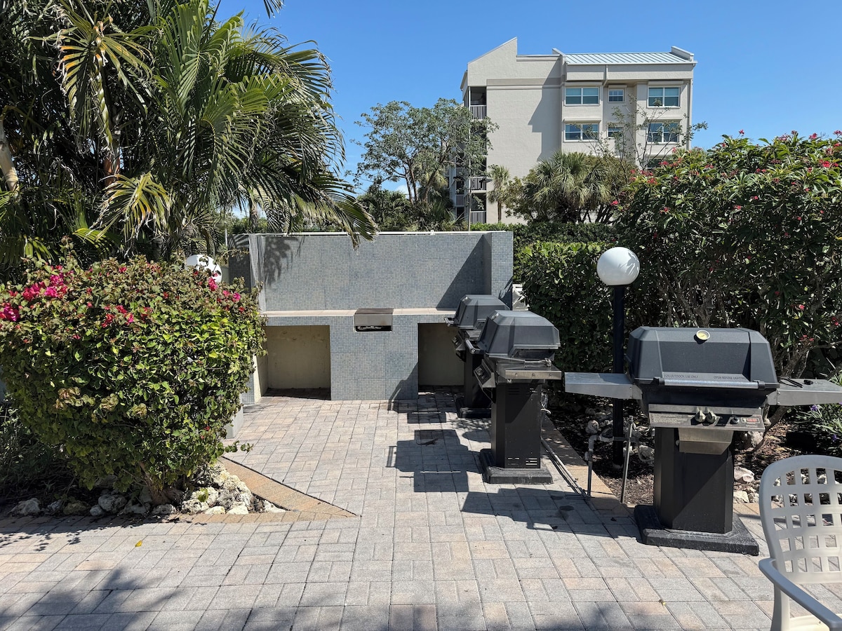 An outdoor patio area with a barbecue setup, a seating area, and a view of a building in the background.