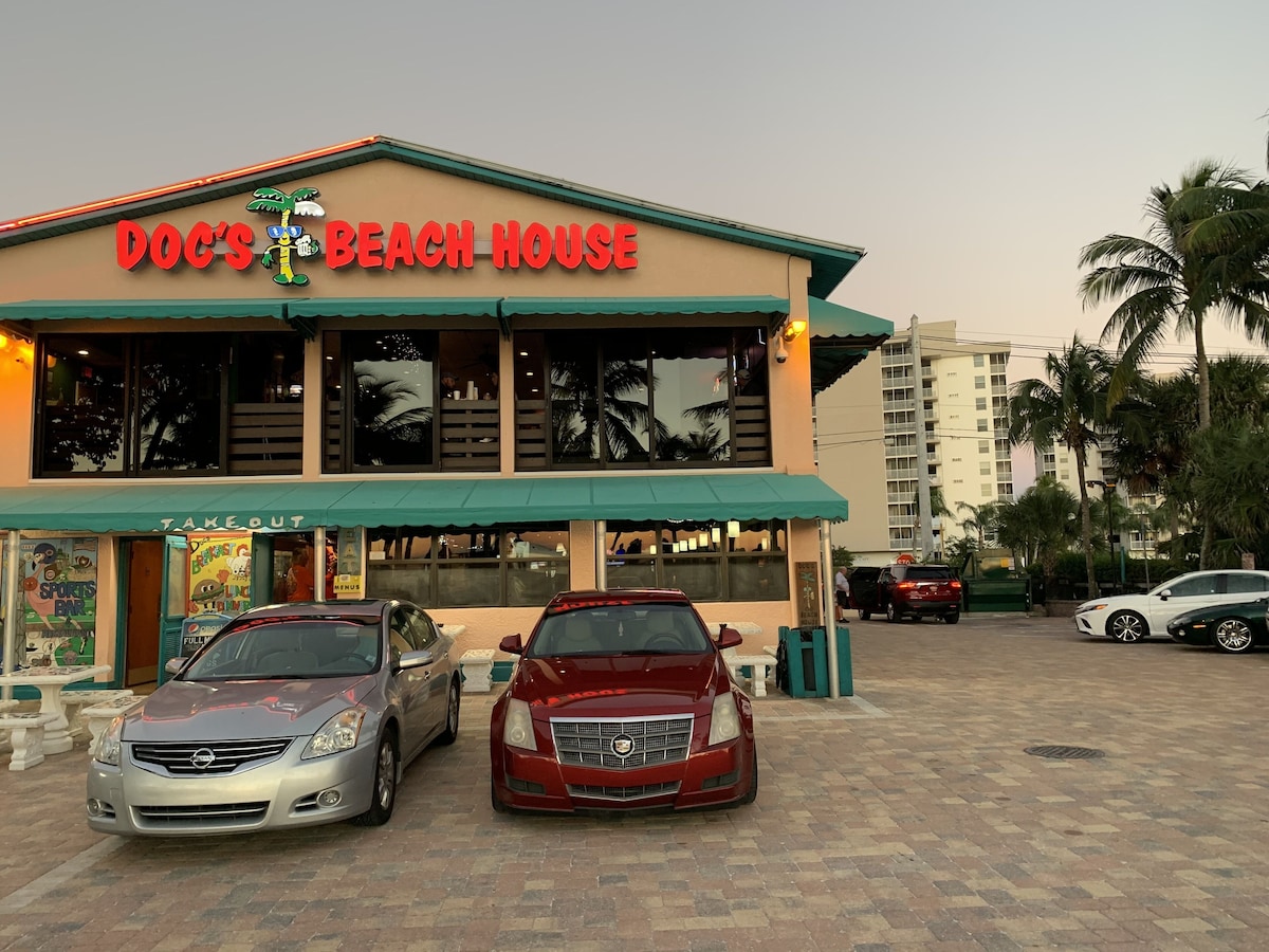 A building with a sign reading Doc's Beach House and a Take Out sign. There are two cars parked in front.
