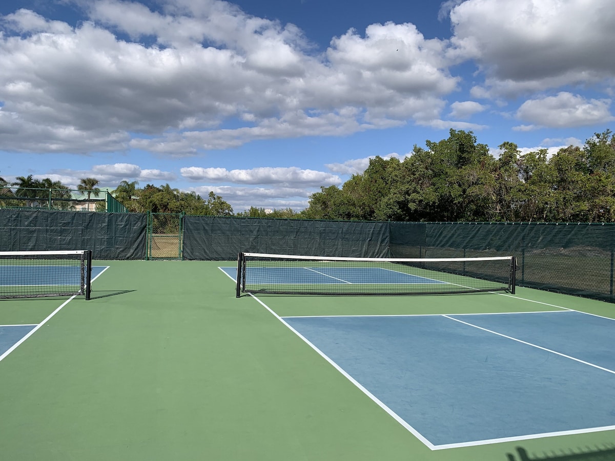 A tennis court with a net, surrounded by a green fence and trees.