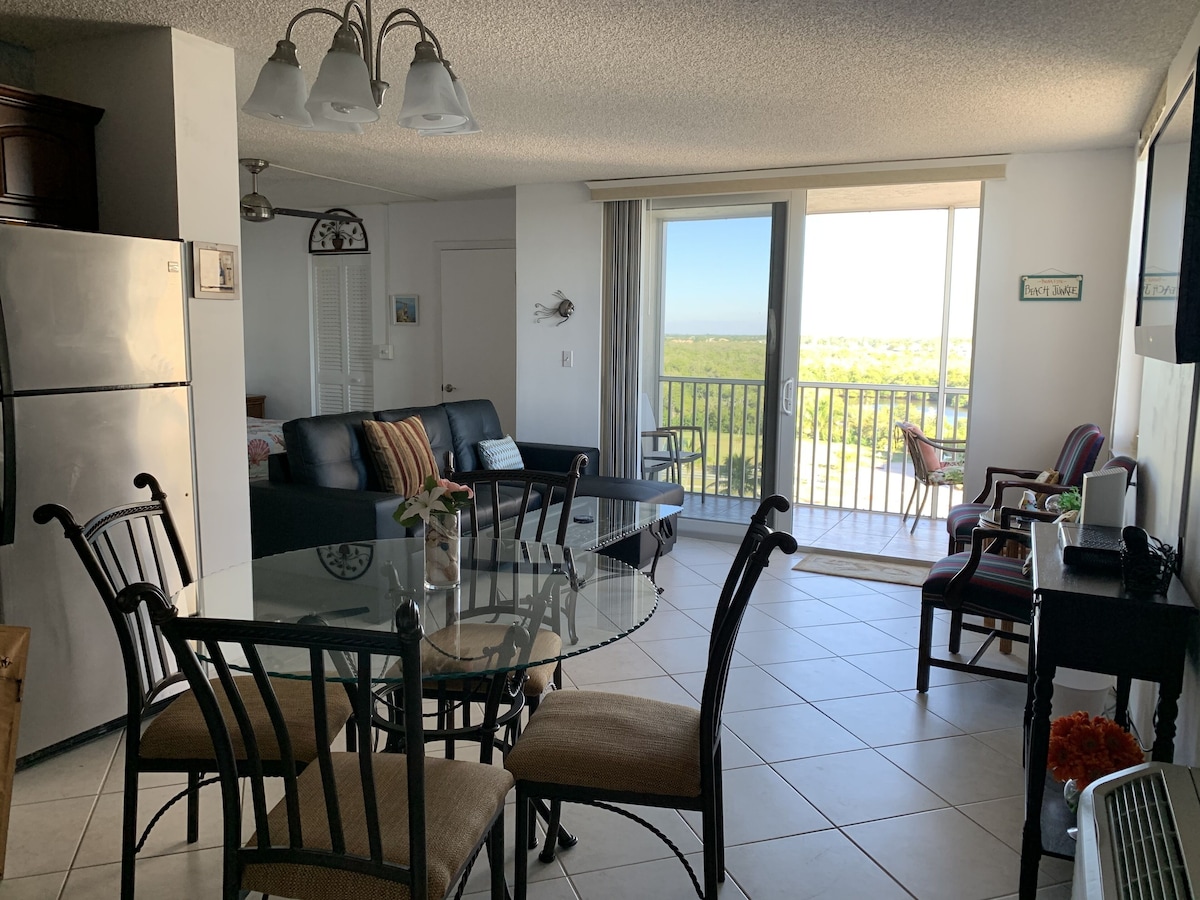 A modern living room with a glass dining table, dark wooden chairs, a black leather sofa, and a view of the outdoors through large sliding glass doors.