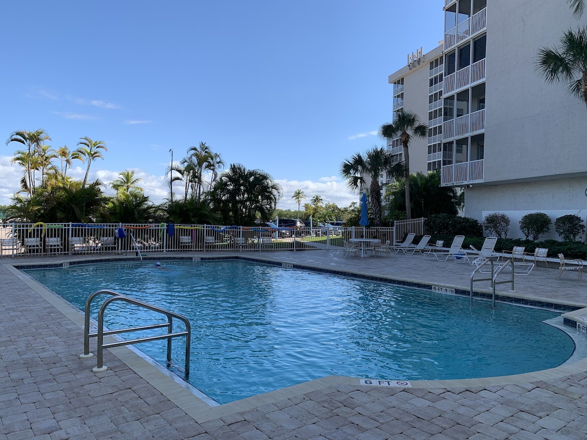 A swimming pool with a 6 FT depth marker, surrounded by lounge chairs and palm trees, with a building in the background.