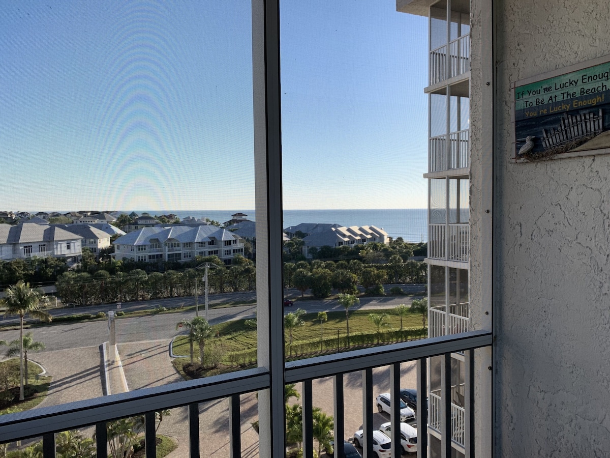 A view from a balcony overlooking a residential area with a road, parked cars, and a building with a sign that reads If you're lucky enough to be at the beach, you're lucky enough!.