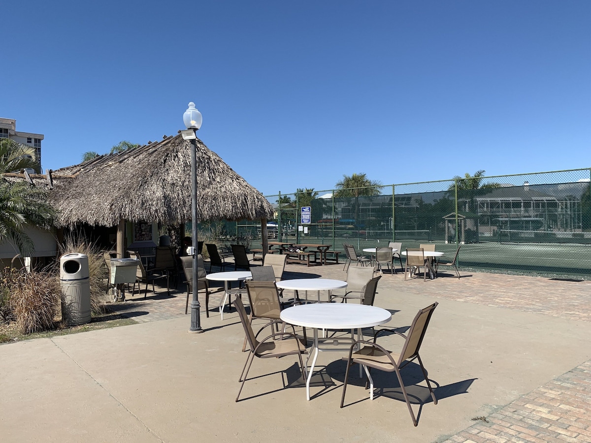 An outdoor patio with round tables and chairs, a thatched-roof structure, and a tennis court in the background.