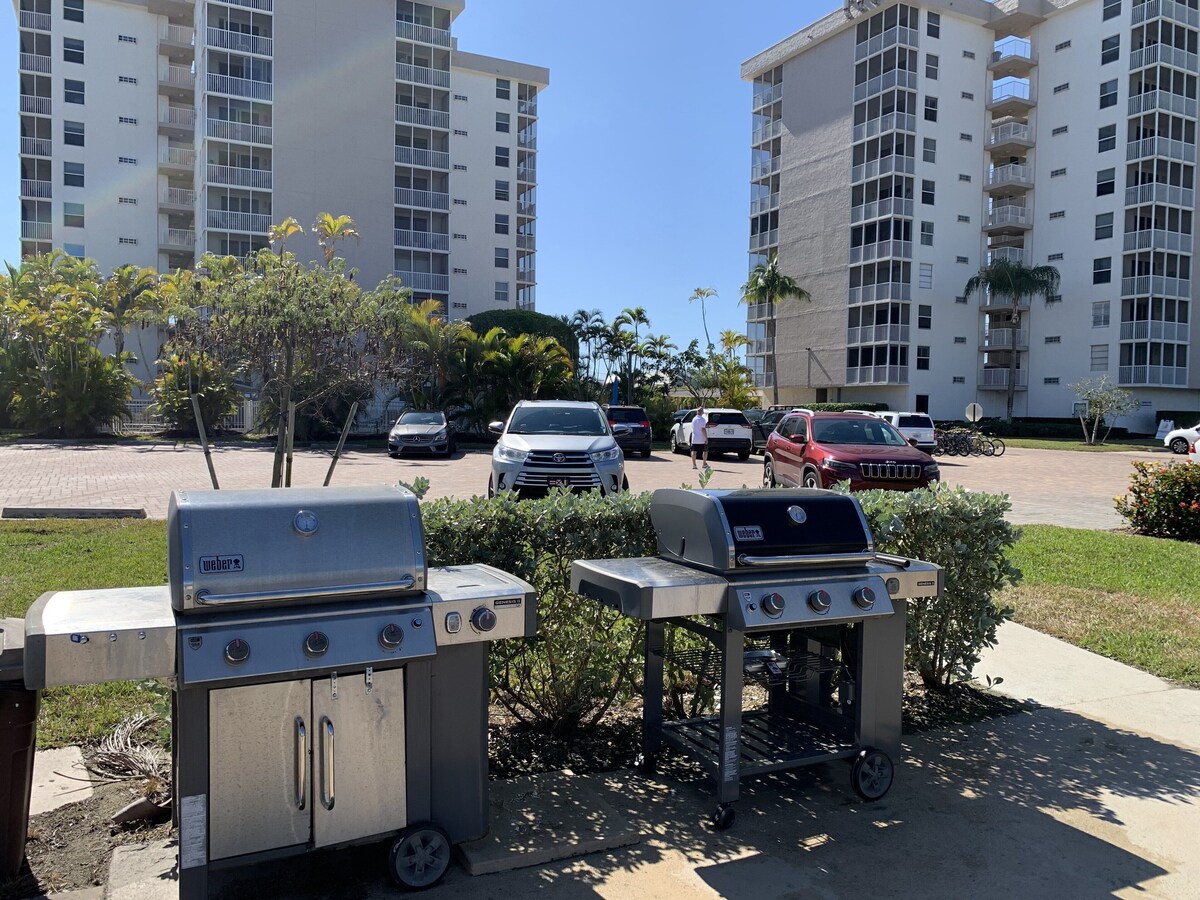 A parking area with two portable grills, a row of parked cars, and two high-rise buildings in the background.