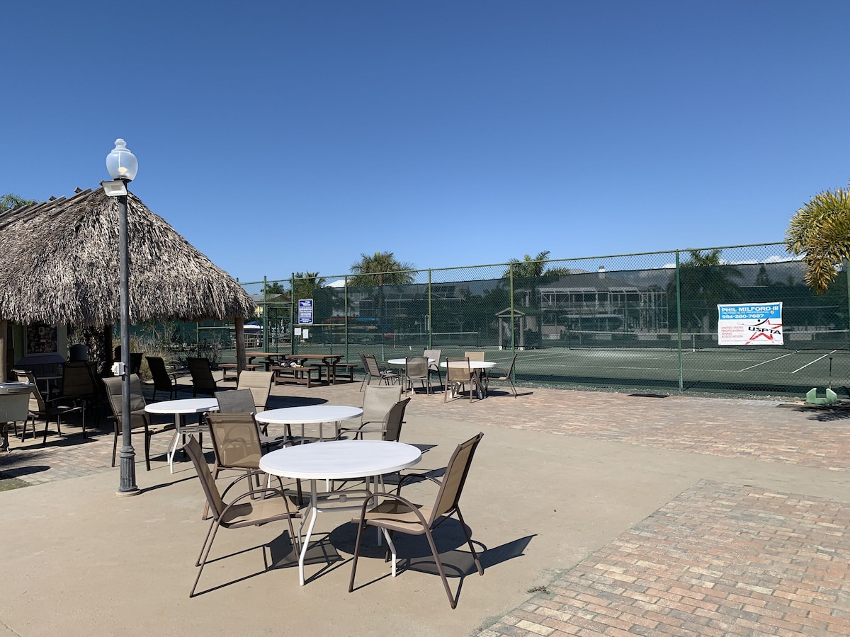 An outdoor seating area with tables and chairs, a thatched umbrella, and a tennis court in the background.