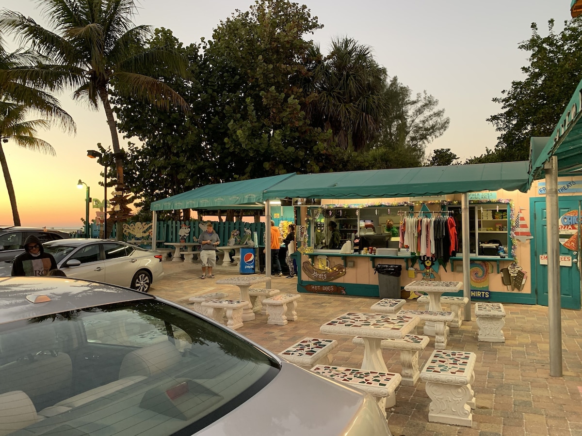 A street food stall with a green awning, seating area with patterned stools, and a car parked nearby.