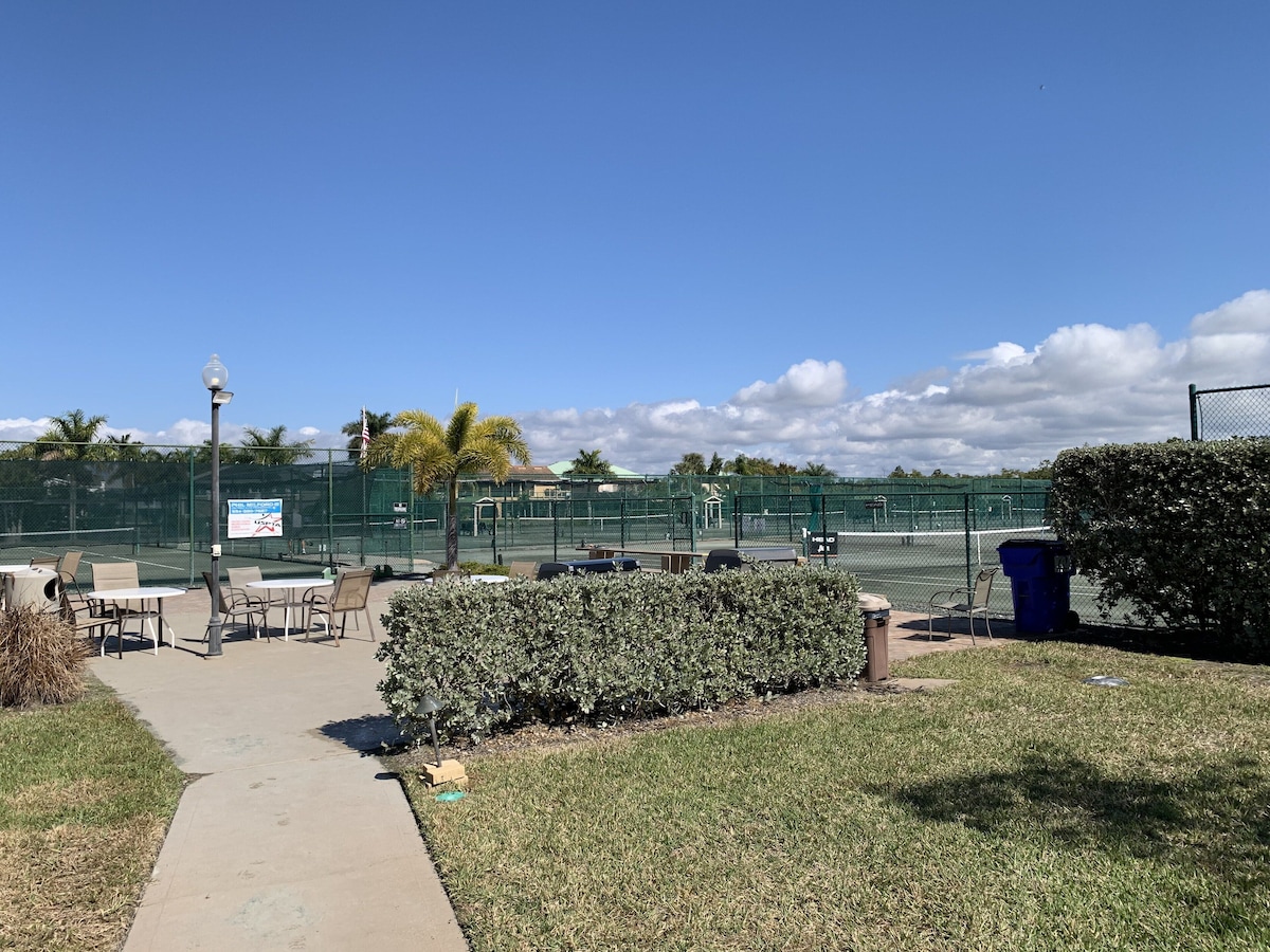 A tennis court with a net, a pathway leading to the court, and a seating area with tables and chairs.