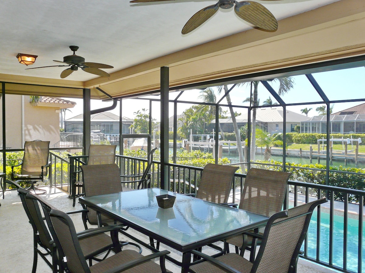 A glass-enclosed patio with a table and chairs, a ceiling fan, and a view of a pool and greenery.