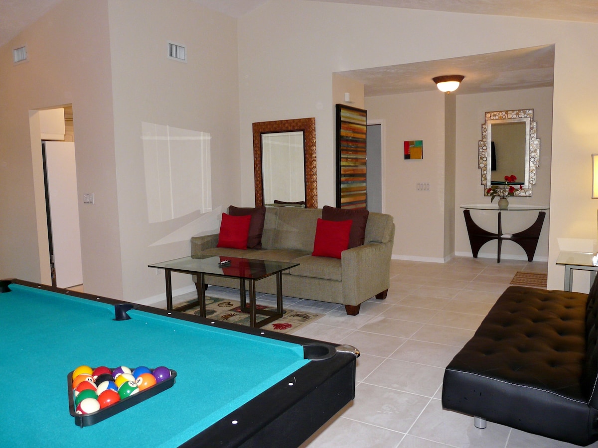 A living room with a pool table, a sofa with red cushions, a coffee table, and a mirror on the wall.