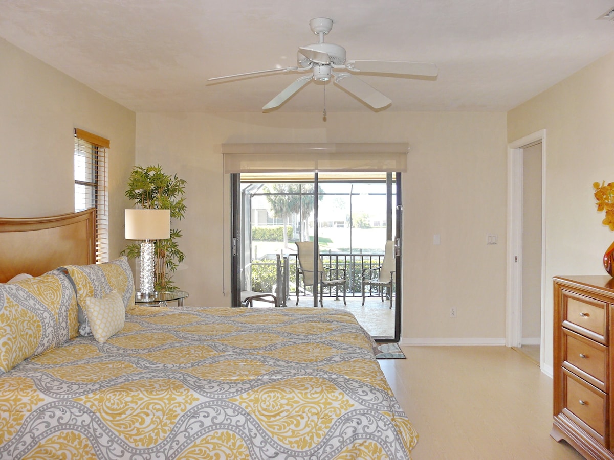 A bedroom with a bed, a ceiling fan, a nightstand, a lamp, and a view of the outside.