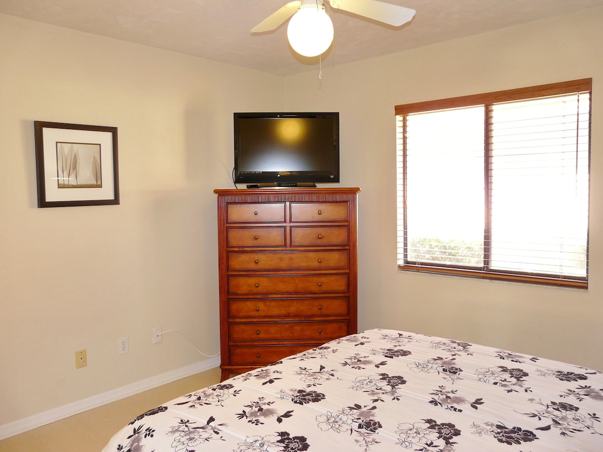 A bedroom with a bed, dresser, television, window, and a ceiling fan.
