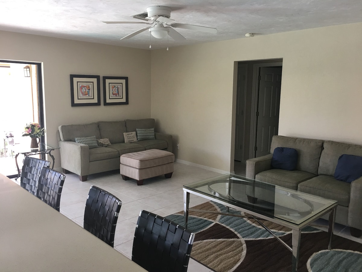 A living room with a glass coffee table, two sofas, a dining table with chairs, and a ceiling fan.