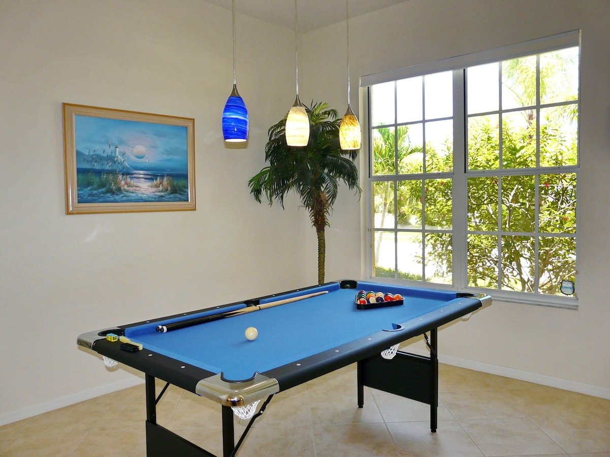 A pool table with balls, a framed painting, and a window with a view of greenery.