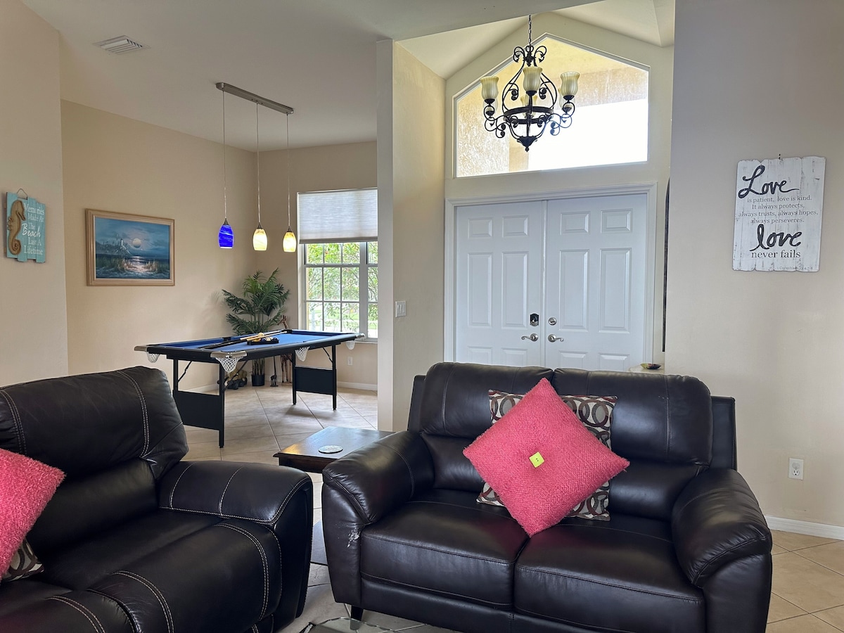 A living room with a pool table, two leather sofas with red cushions, a chandelier, and a door with a 'Love never fails' sign.