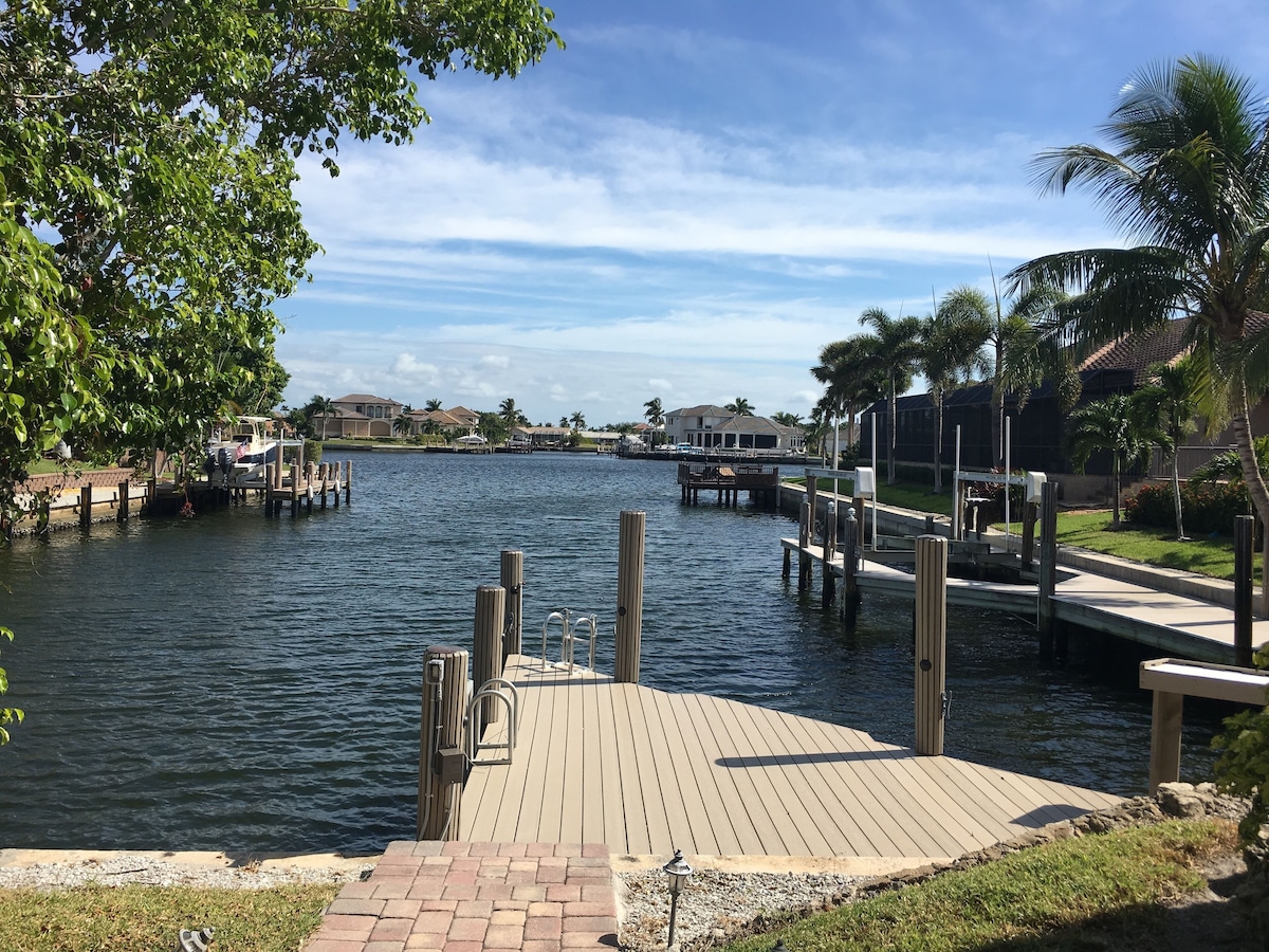 A waterfront dock with a wooden walkway leading into calm water, surrounded by greenery and residential buildings.