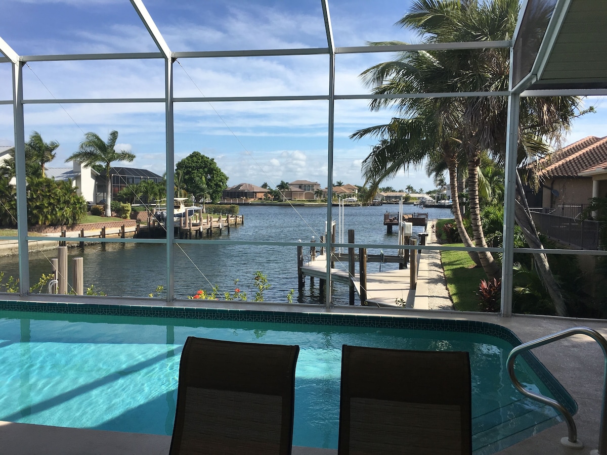 A pool with two chairs, a view of a marina with boats, and palm trees.