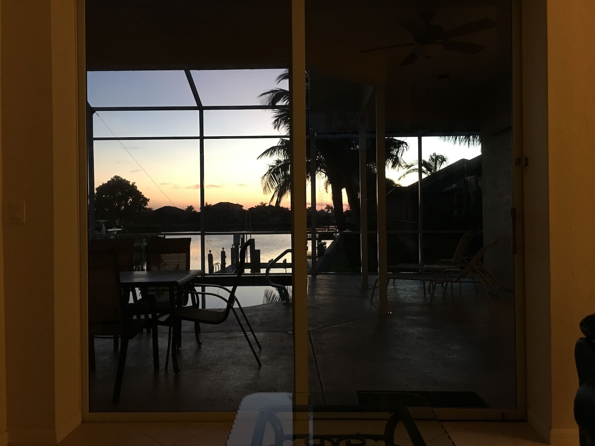 A room with a large sliding glass door leading to an outdoor area with a pool and chairs, and a view of the sunset over the water.