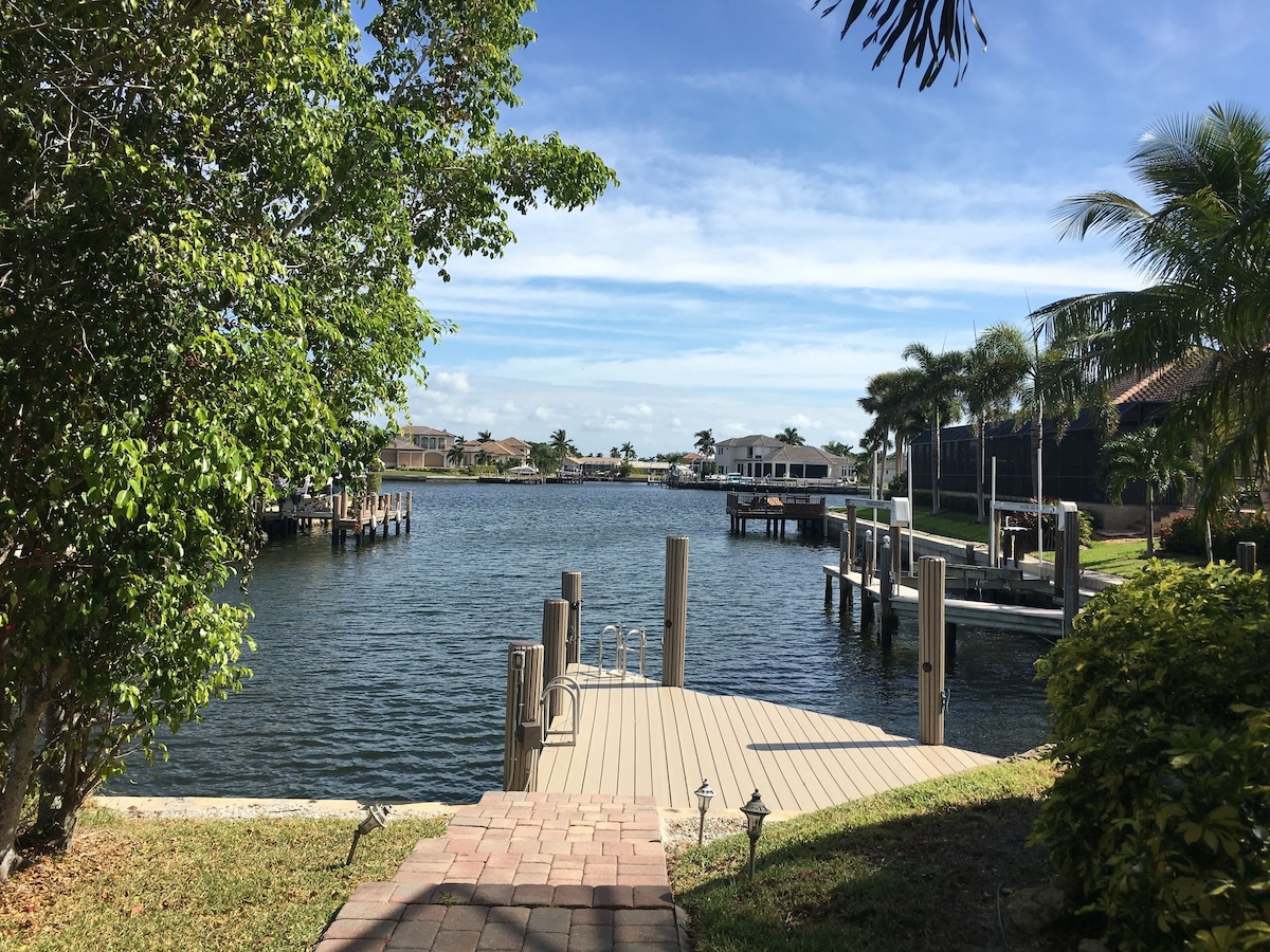 A waterfront property with a dock extending into a calm body of water, surrounded by lush greenery and residential buildings in the background.
