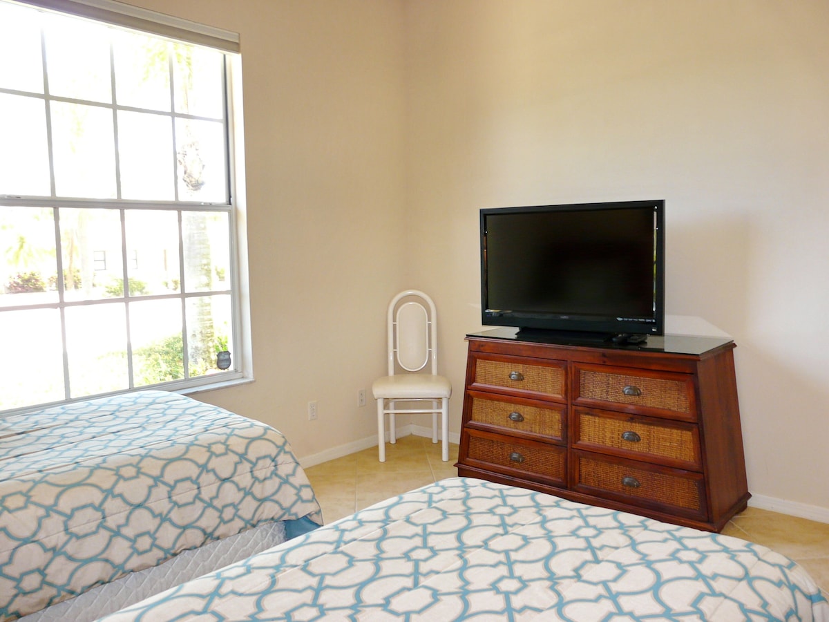 A bedroom with two beds, a television on a dresser, a chair, and a window with a view of greenery.