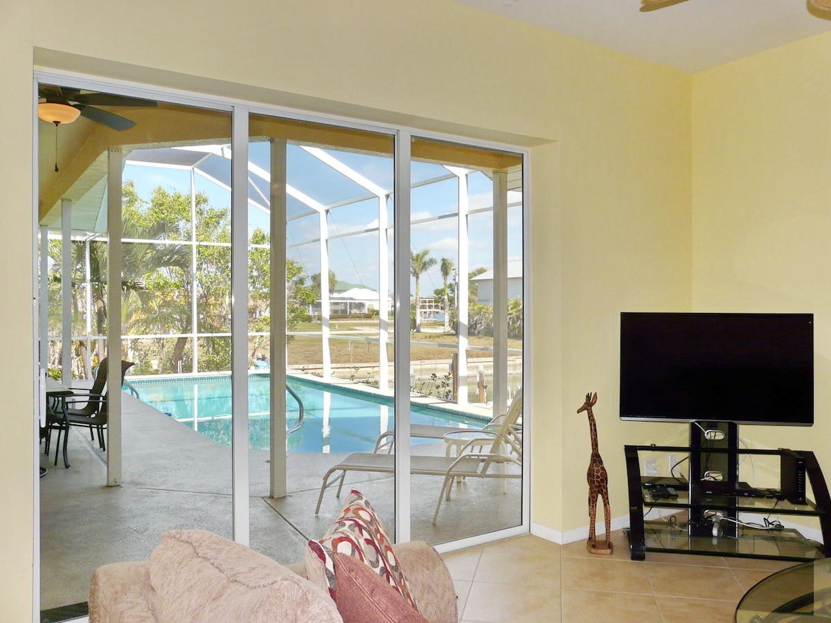 A living room with a large sliding glass door leading to a pool area, a television on a stand, and a giraffe statue.