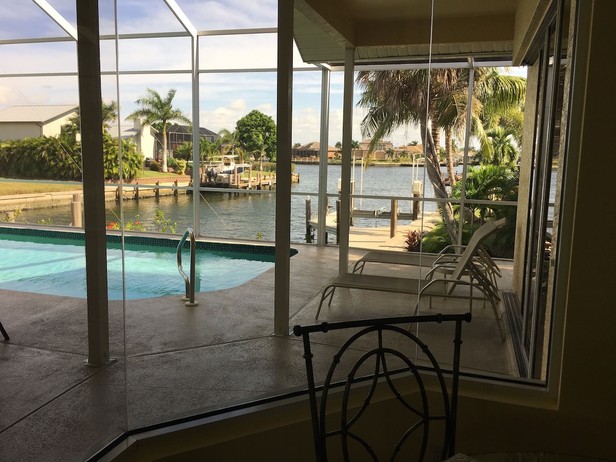 A pool area with a view of a body of water, palm trees, and a covered seating area.