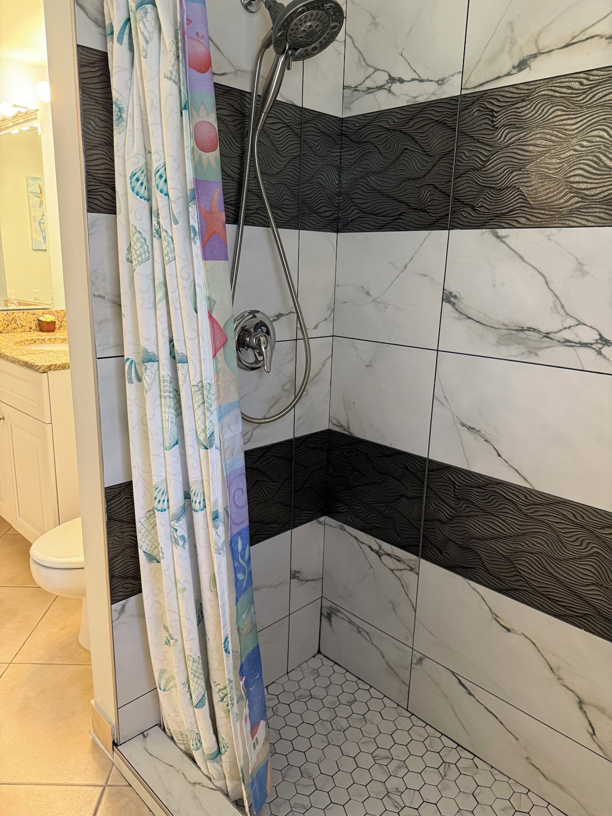 A shower with a handheld showerhead, a curtain with a fish pattern, and hexagonal tiles on the floor.