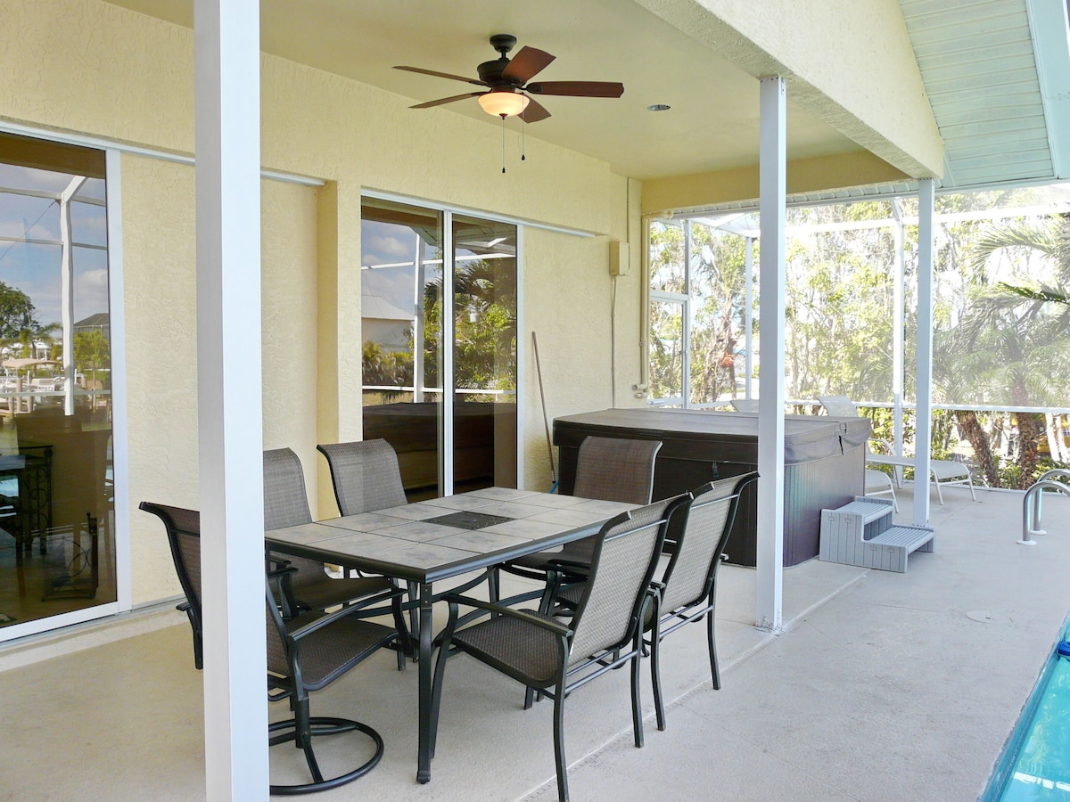 A covered patio with a table and chairs, a ceiling fan, and a view of the pool and greenery.