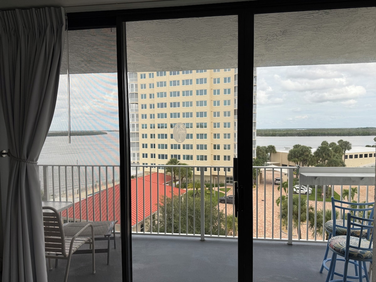 From inside the slider, a tall beige tower with a palm emblem blocks the center, with the bay to the left and right, white balcony chairs and a table, and blue bar stools