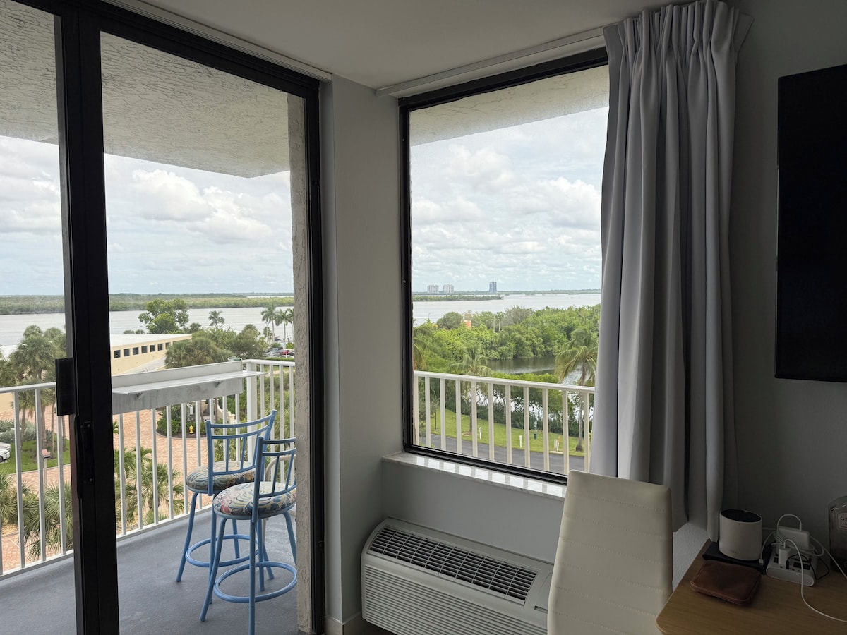 Balcony door and a window over a wide bay, mangroves, and distant towers, with blue balcony chairs
